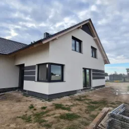 Nowoczesny dom jednorodzinny z białą elewacją, czarnymi akcentami i oknami, widok z zewnątrz na plac budowy z rusztowaniami i otoczeniem w trakcie zagospodarowywania.