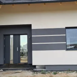 Nowoczesna elewacja domu jednorodzinnego z grafitowymi pasami, szarą stolarką okienną i drzwiami, widoczne prace wykończeniowe wokół budynku.