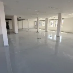 Przestronne, jasne wnętrze biurowe z szarym, błyszczącym wykończeniem podłogi, białymi ścianami i kolumnami. Aluminiowa drabina z wiadrem i pędzlem stoi przy oknie, sugerując prace wykończeniowe.