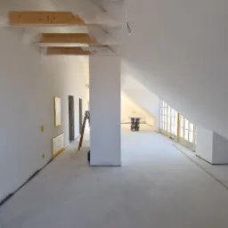 Przestronne, jasne poddasze w trakcie wykończenia, z białymi ścianami i sufitami, zabezpieczonymi drewnianymi belkami stropowymi, widoczne drabina, puste otwory drzwiowe i okna zakryte folią...