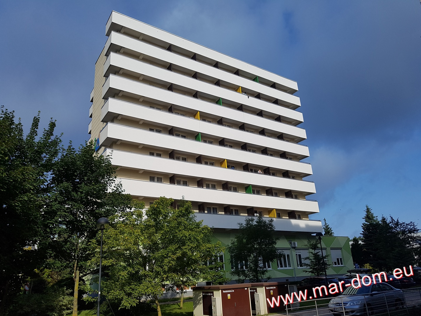 Wysoki budynek mieszkalny z białymi balkonami, widok z dołu, częściowo zasłonięty zielenią drzew