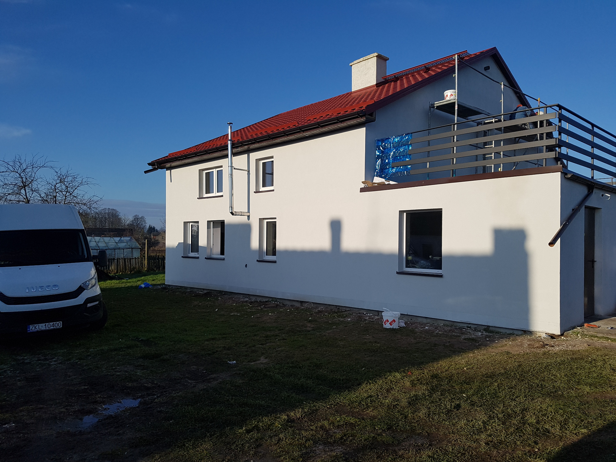 Dwupiętrowy dom z nową, białą elewacją i czerwoną dachówką, widok z boku na tle błękitnego nieba, z zaparkowanym białym samochodem dostawczym i widocznym rusztowaniem na balkonie.