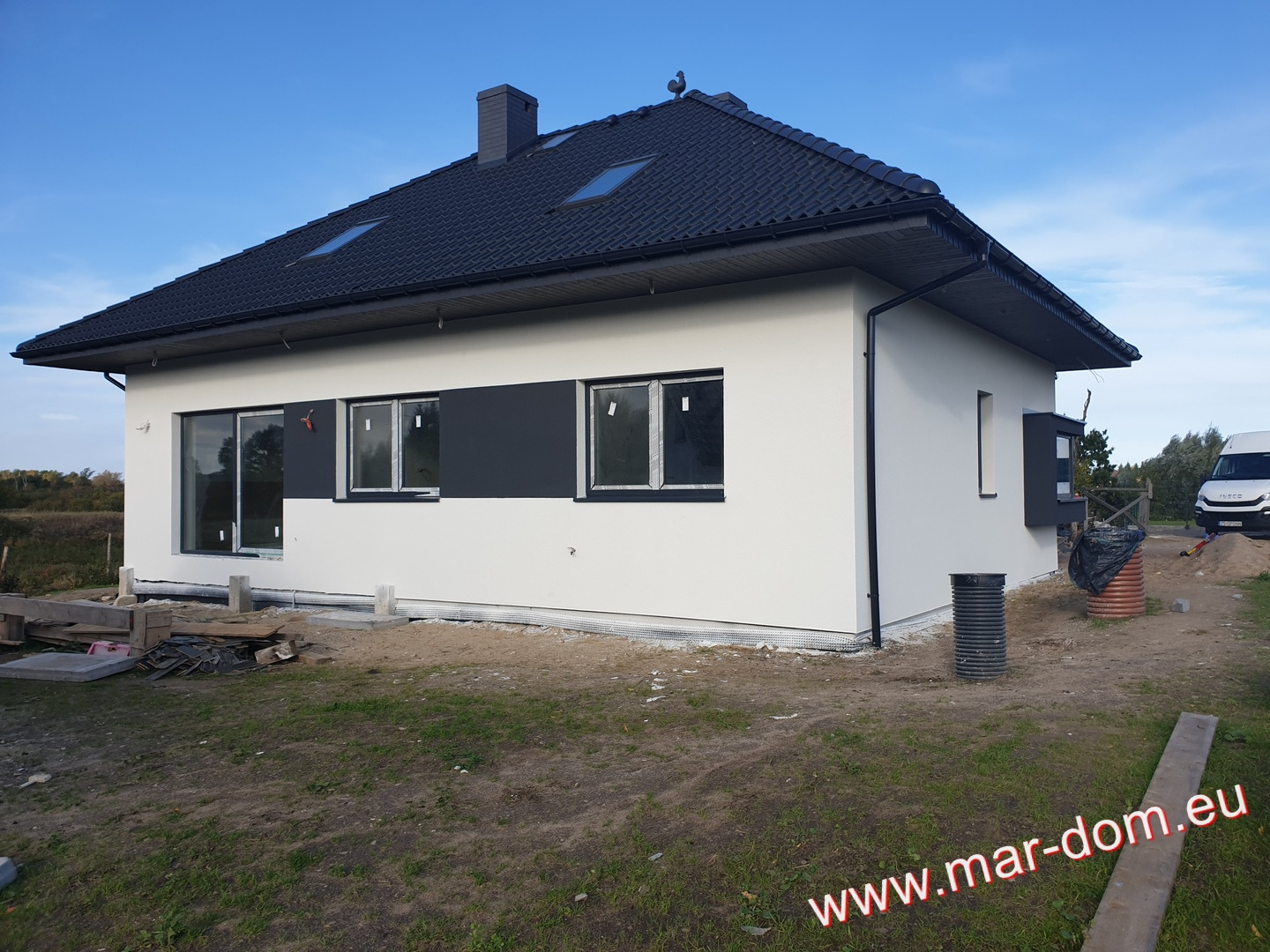 Nowoczesny dom jednorodzinny z białą elewacją i czarnym dachem, widoczne czarne obramowania okien i czarny fragment elewacji, teren wokół domu częściowo zagospodarowany, widoczne materiały budowlane.