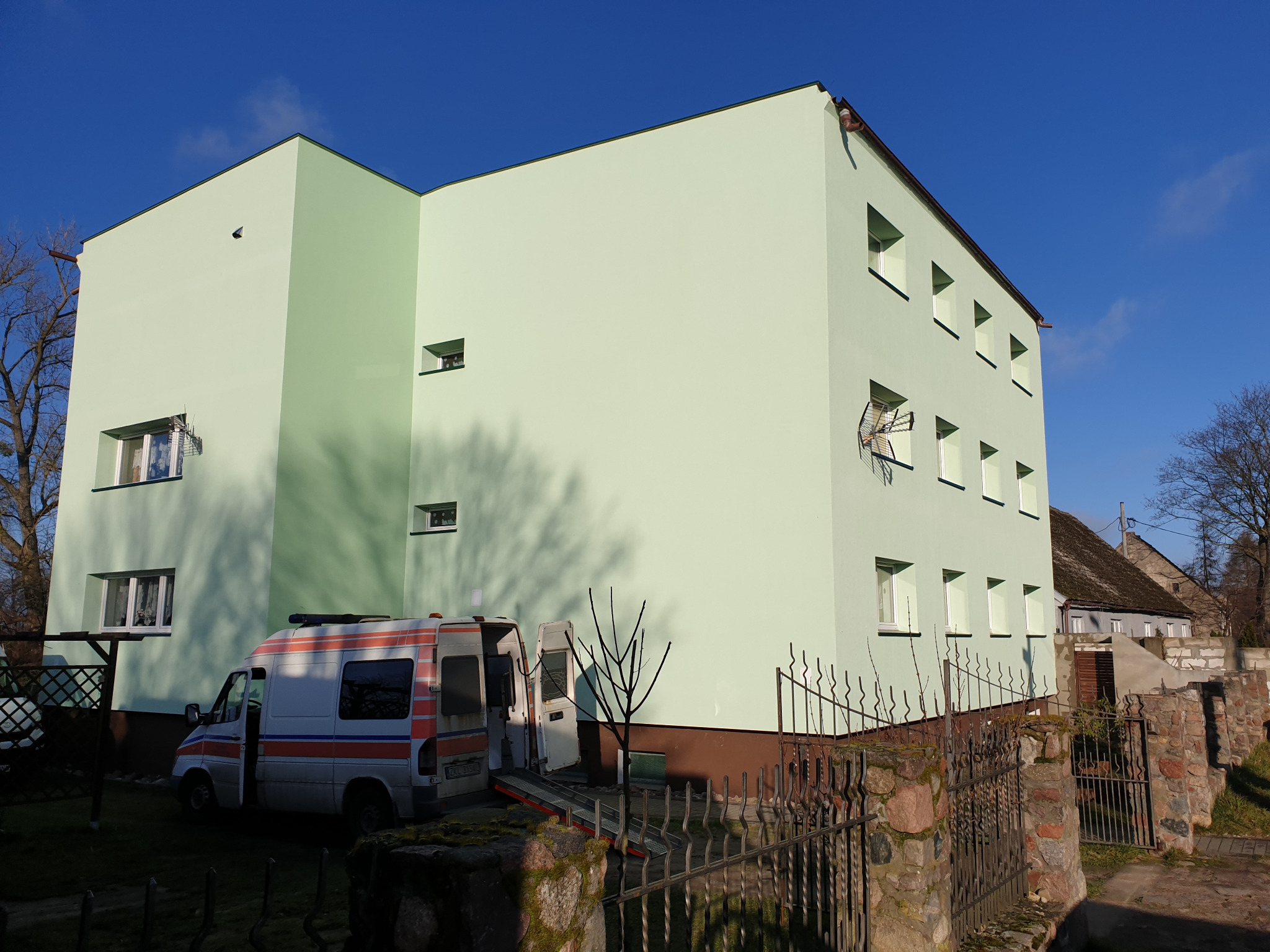 Dwukondygnacyjny budynek po renowacji elewacji w odcieniach zieleni, z zaparkowaną karetką pogotowia w pobliżu, widok zza metalowego ogrodzenia.