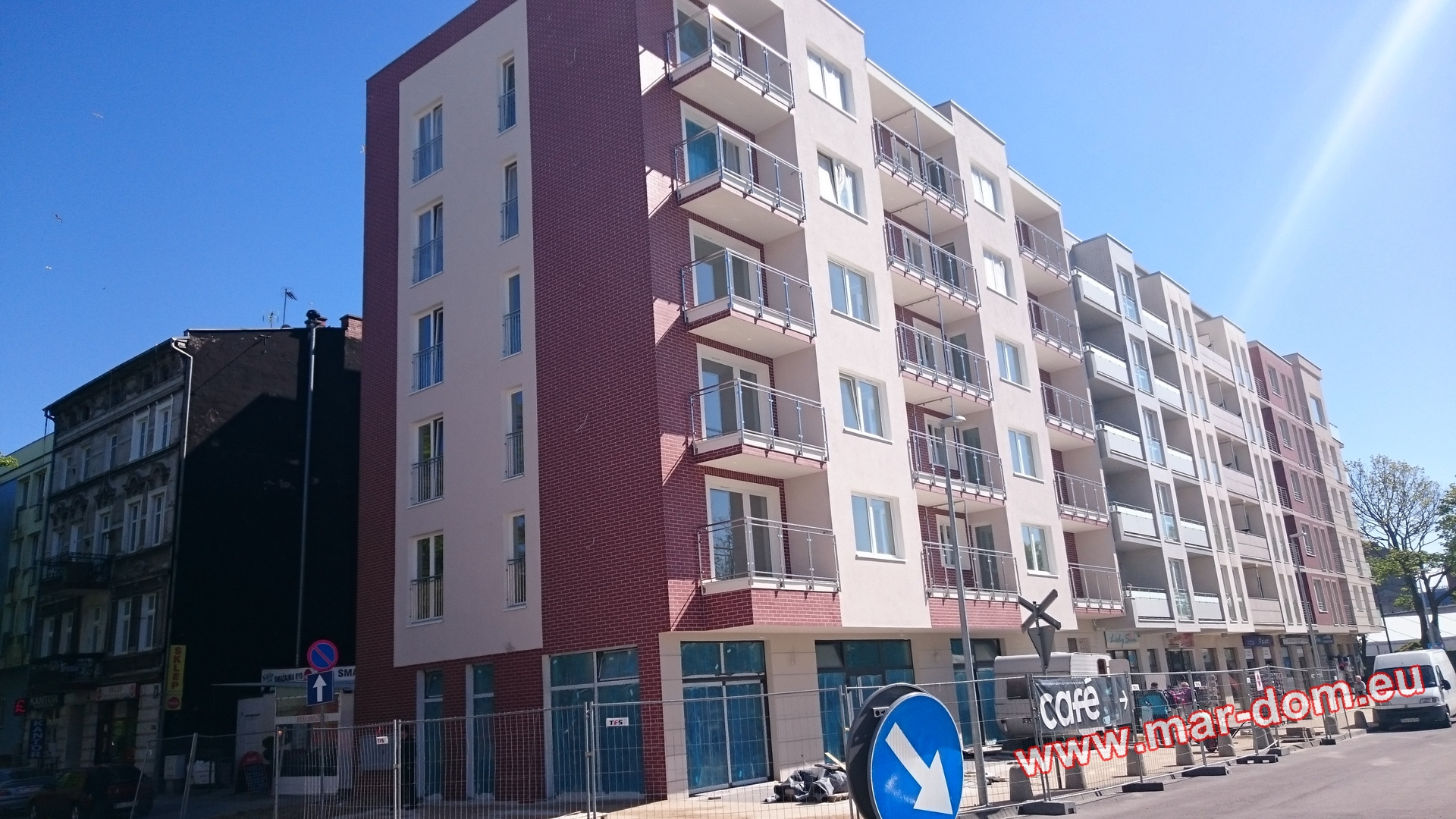 Nowo wybudowany, wielopiętrowy budynek mieszkalny z balkonami, wykończony w jasnych kolorach z elementami cegły na elewacji, widoczny z poziomu ulicy, z tymczasowym ogrodzeniem budowlanym.