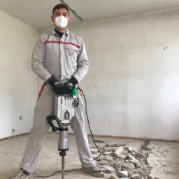 Pracownik w kombinezonie i masce FFP2, z młotem udarowym Blank & Hagen, stoi na gruzowisku w pomieszczeniu z surowymi ścianami.