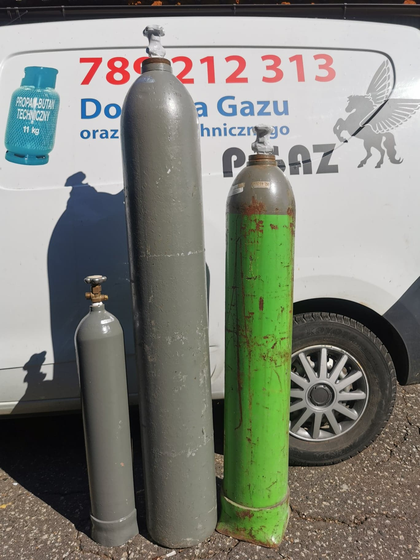 Trzy butle z gazem: mała szara z zaworem, wysoka szara i zielona z widocznymi śladami użytkowania, na tle samochodu dostawczego z numerem telefonu i logo firmy.