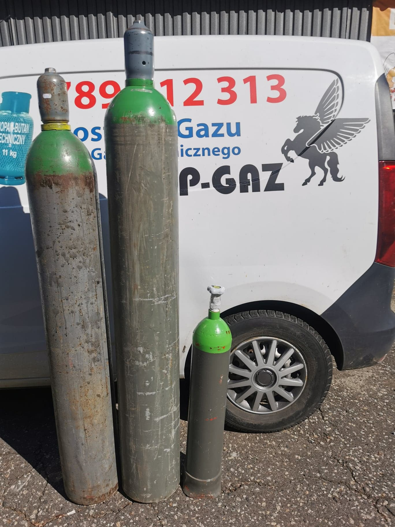 Butle z gazem o różnej wielkości i stopniu zużycia, ustawione przed białym samochodem dostawczym z logo firmy P-GAZ.