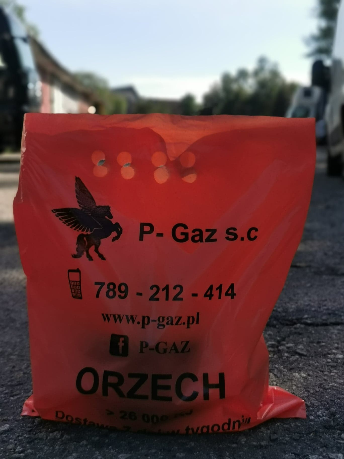 Pomarańczowa torba reklamowa firmy P-Gaz s.c. z logo pegaza, numerem telefonu i adresem strony internetowej, leżąca na asfaltowej nawierzchni.
