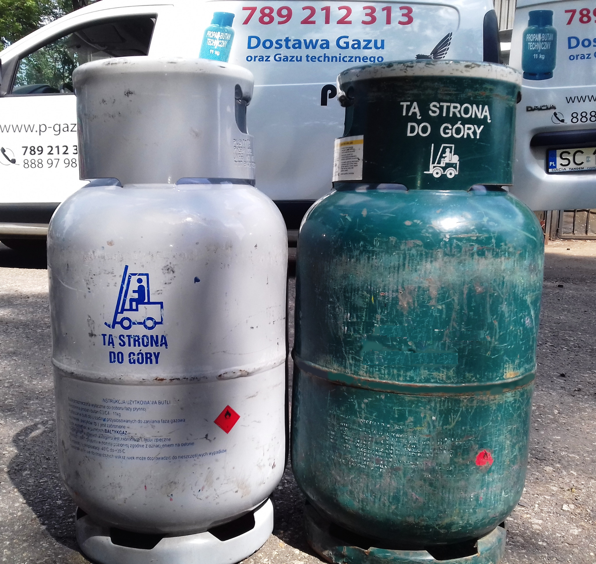 Dwie butle gazowe, biała i zielona, z naklejkami informacyjnymi i symbolem wózka widłowego, stojące na zewnątrz, z dostawczym samochodem z reklamą dostawy gazu w tle.