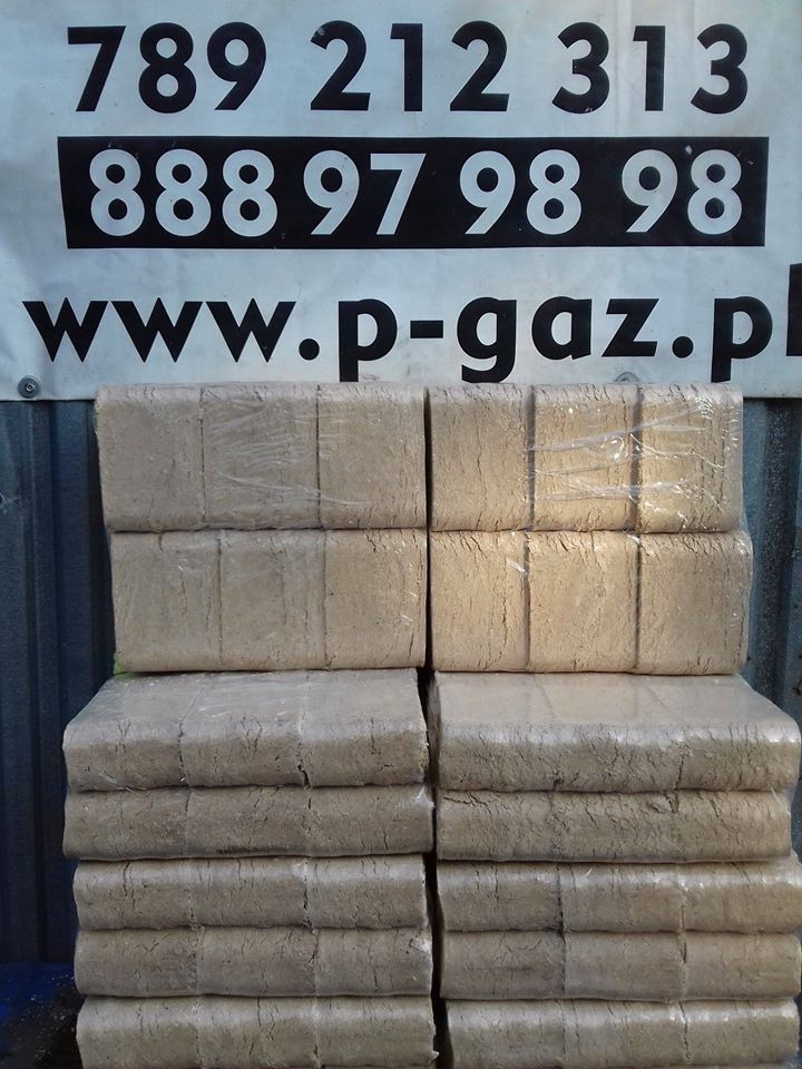 Ułożone w stosy pakiety brykietu drzewnego owinięte folią, na tle baneru reklamowego z numerami telefonów i adresem strony internetowej.