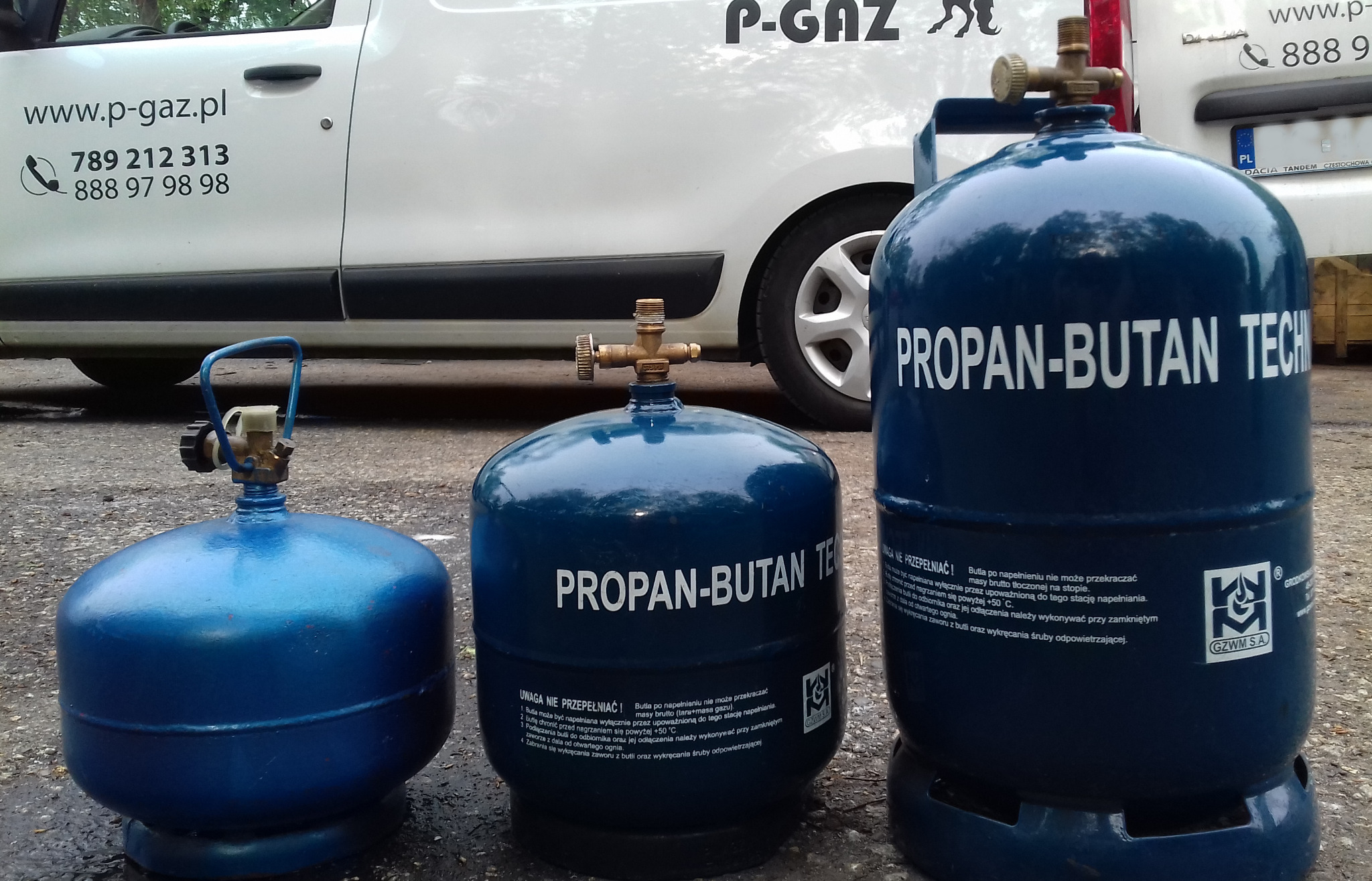 Trzy butle z gazem propan-butan w różnych rozmiarach, ustawione przed białym samochodem dostawczym z logo i numerami kontaktowymi firmy.