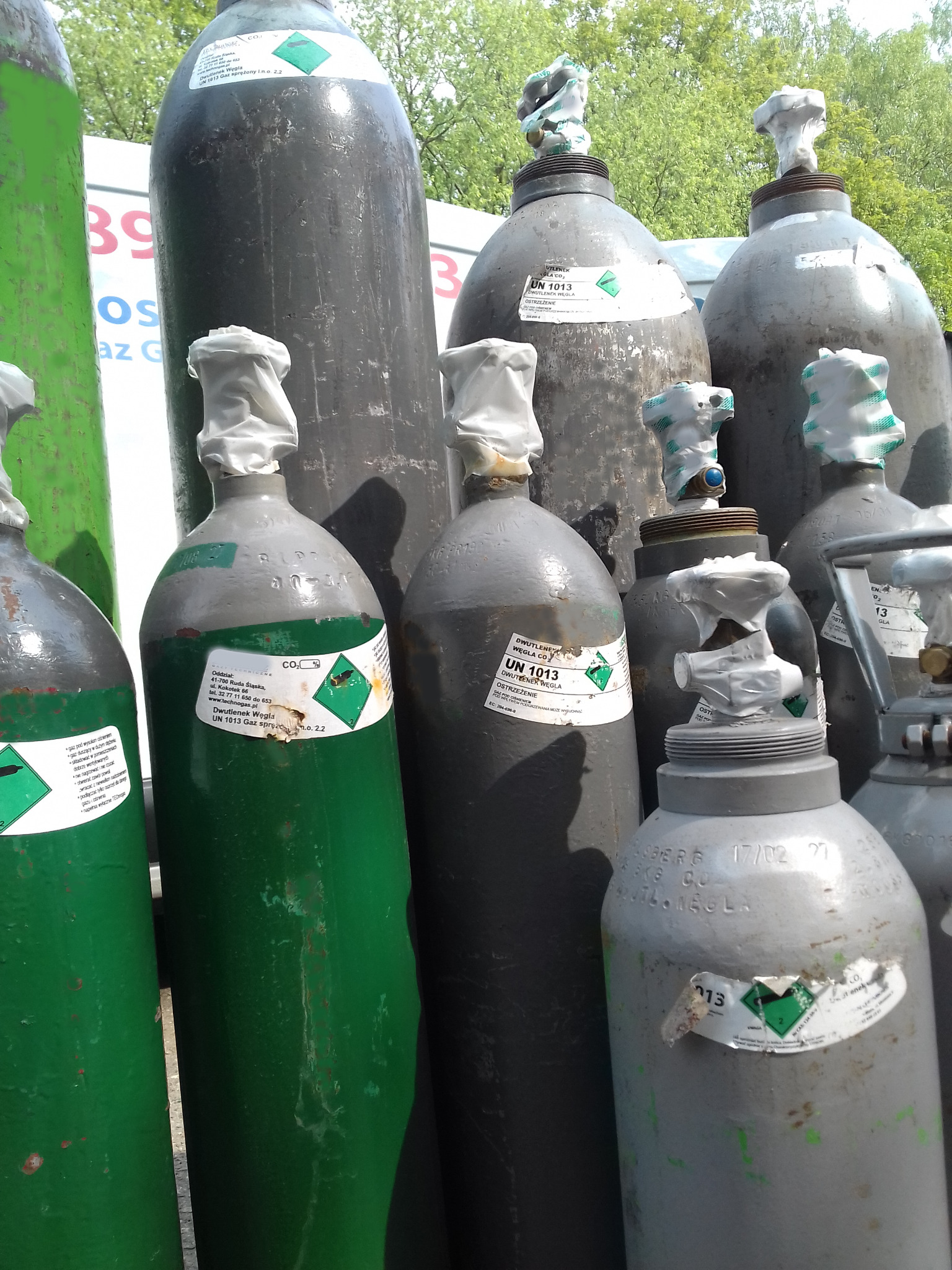 Grupa butli z dwutlenkiem węgla w kolorach zielonym i szarym, z widocznymi etykietami ostrzegawczymi i zabezpieczeniami na zaworach, ustawione na zewnątrz w słoneczny dzień.