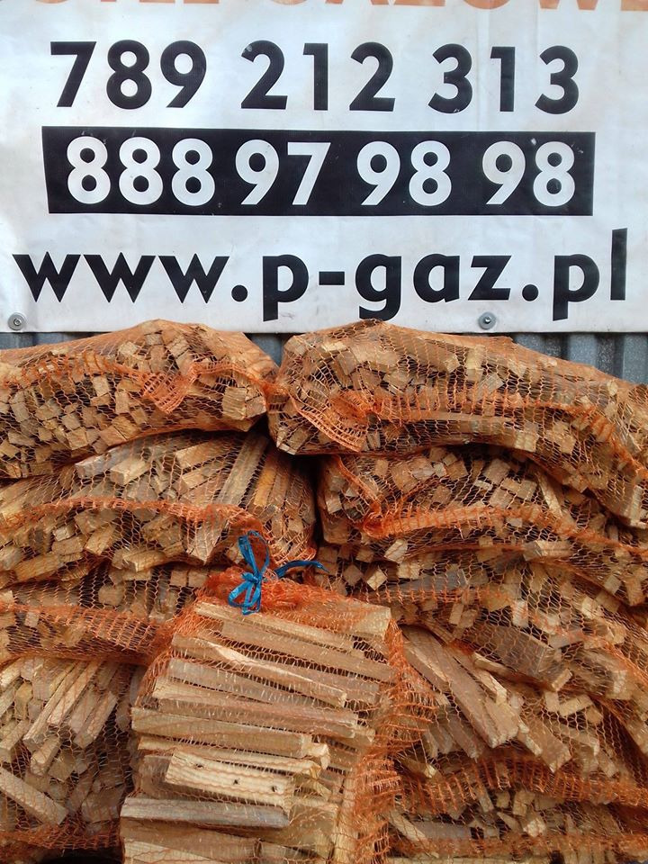 Stosy worków z pociętym drewnem kominkowym w pomarańczowej siatce na tle baneru reklamowego z numerami telefonów i adresem strony internetowej