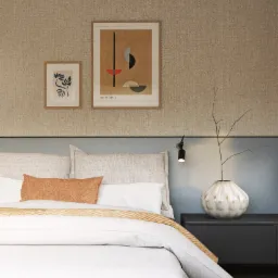 Nowoczesna sypialnia z łóżkiem z szarym zagłówkiem, pomarańczową poduszką, dwiema grafikami na ścianie i wazonem z gałązkami na szafce nocnej.