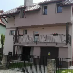 Dwupiętrowy dom z beżową elewacją, balkonem z ozdobną balustradą i czerwonymi kwiatami w donicach, widok zza czarnego ogrodzenia z ceglanymi słupkami.