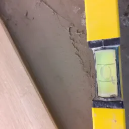 Żółta poziomica przyłożona do świeżo nałożonej, szarej warstwy zaprawy na ścianie, obok ułożone panele podłogowe.