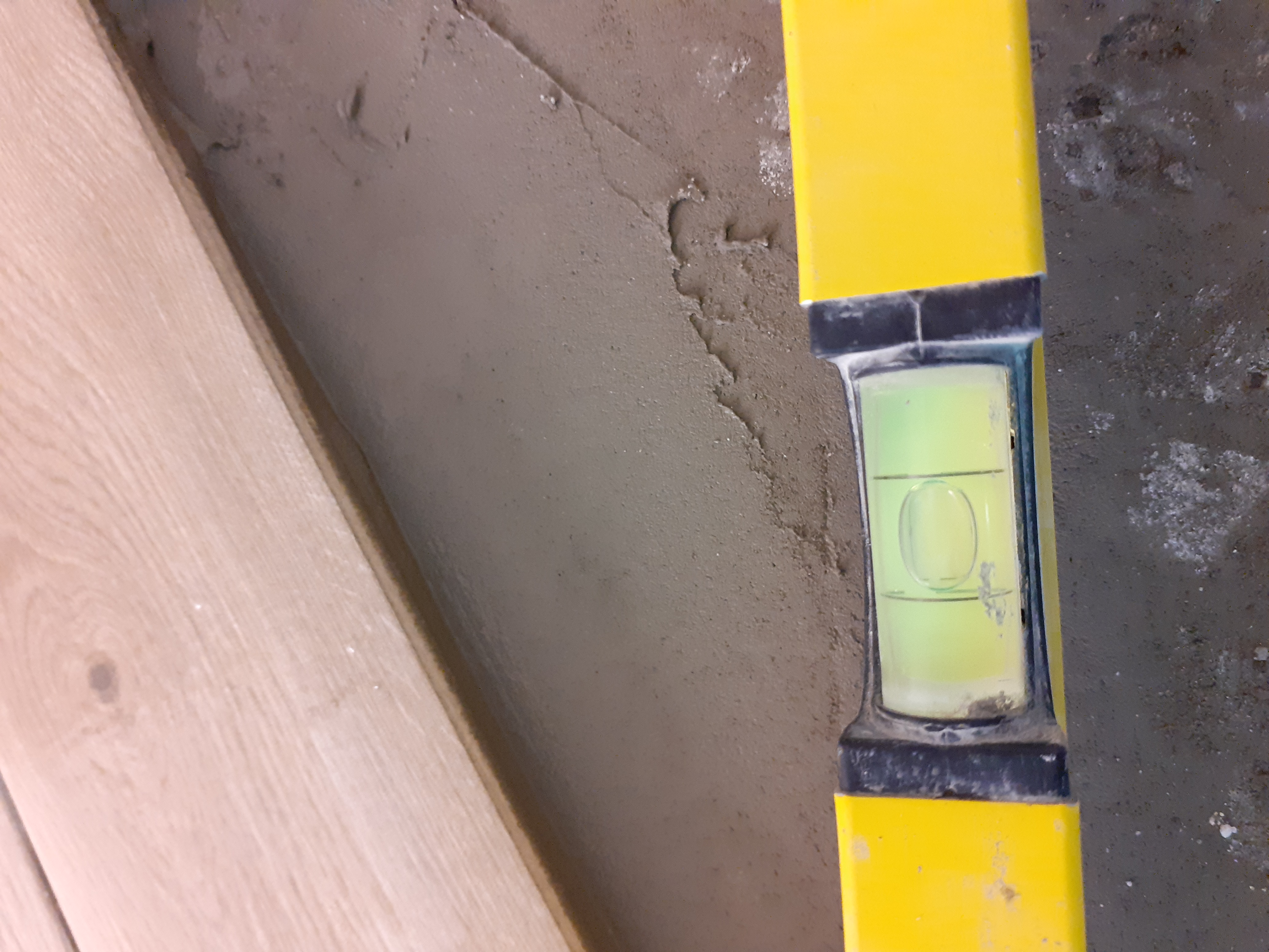Żółta poziomica przyłożona do świeżo nałożonej, szarej warstwy zaprawy na ścianie, obok ułożone panele podłogowe.