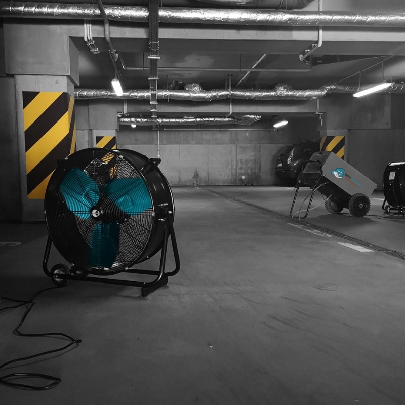 Duży wentylator z niebieskimi łopatkami na tle szarego parkingu podziemnego w Łodzi, z żółto-czarnymi pasami na kolumnach i innymi urządzeniami do osuszania w tle.