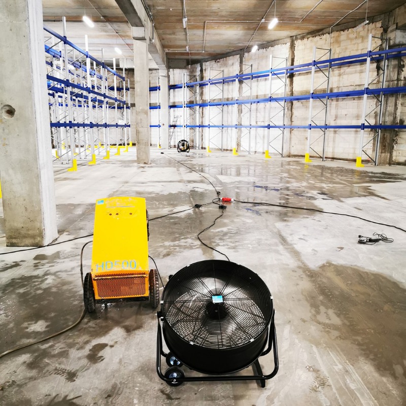 Żółty osuszacz HD500 z wentylatorem na mokrej betonowej podłodze w hali magazynowej z metalowymi regałami. Widoczne kable zasilające i słupy konstrukcyjne.