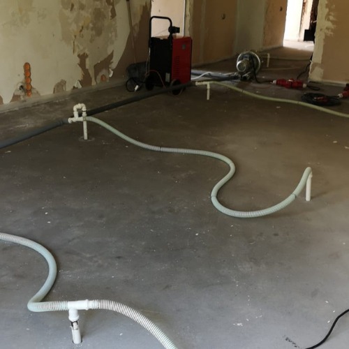 Wnętrze w trakcie prac, widoczne rury i sprzęt do usuwania wilgoci z posadzki, ściany częściowo zdarta farba, widoczne ślady po instalacji.