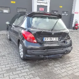 Uszkodzony tył czarnego samochodu Peugeot 207 z polską tablicą rejestracyjną KR 8KF30, widoczne wgniecenia karoserii, pęknięty zderzak i uszkodzone światło, na tle szarej ściany budynku z napisem...