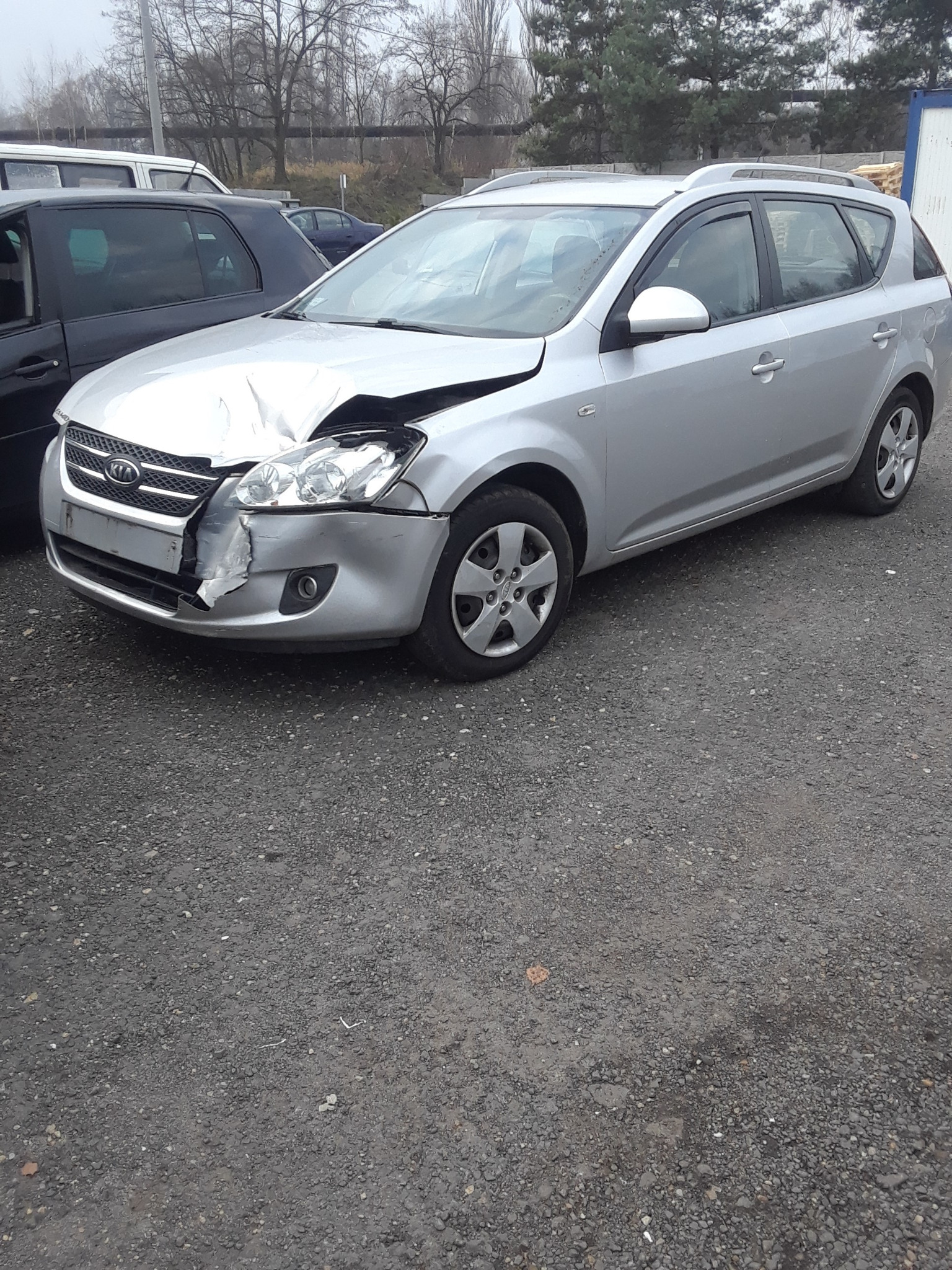 Srebrny samochód kombi marki Kia z uszkodzonym przodem, wgnieciony błotnik i zniszczony reflektor, zaparkowany na szarym żwirowym podłożu.