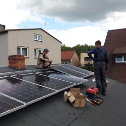 Eco-Fox - Montaż paneli słonecznych na dachu domu jednorodzinnego przez dwóch instalatorów. Widoczna skrzynka z narzędziami, kartony po panelach oraz fragmenty innych budynków w tle.