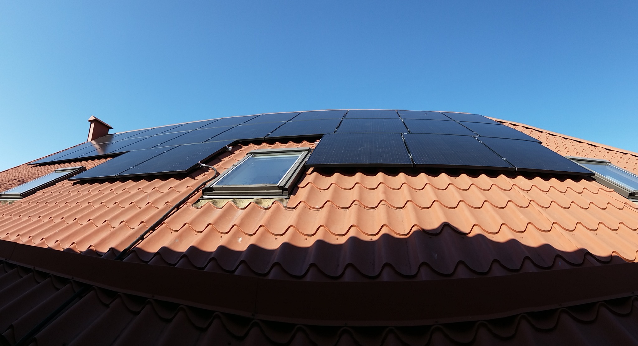 Instalacja paneli fotowoltaicznych na dachu z dachówką ceramiczną, widoczne okna dachowe i przewody, błękitne niebo w tle.