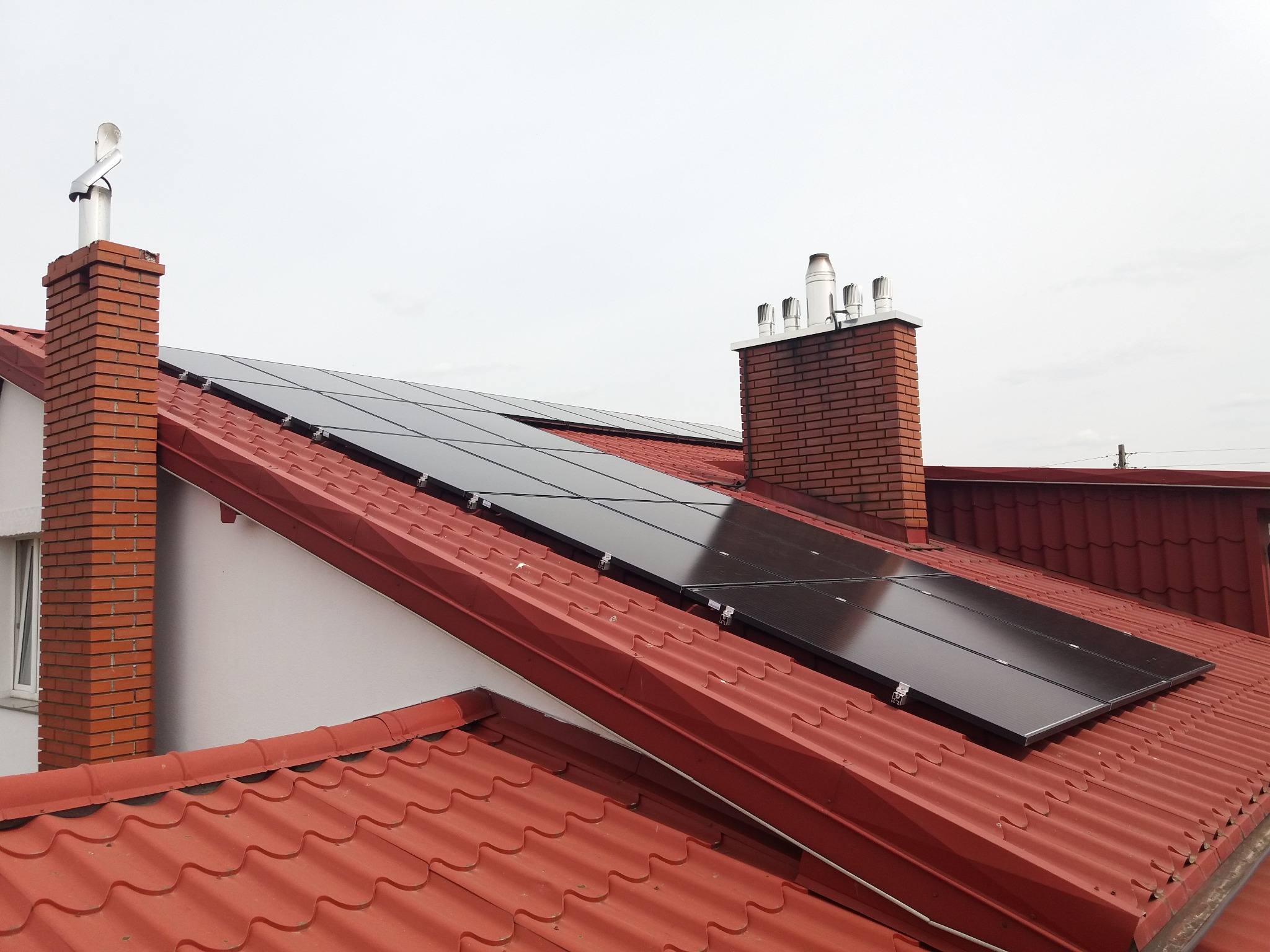 Panele słoneczne 6kW zamontowane na dachu z czerwonej dachówki, widoczny komin z cegły i metalowa rura wentylacyjna.