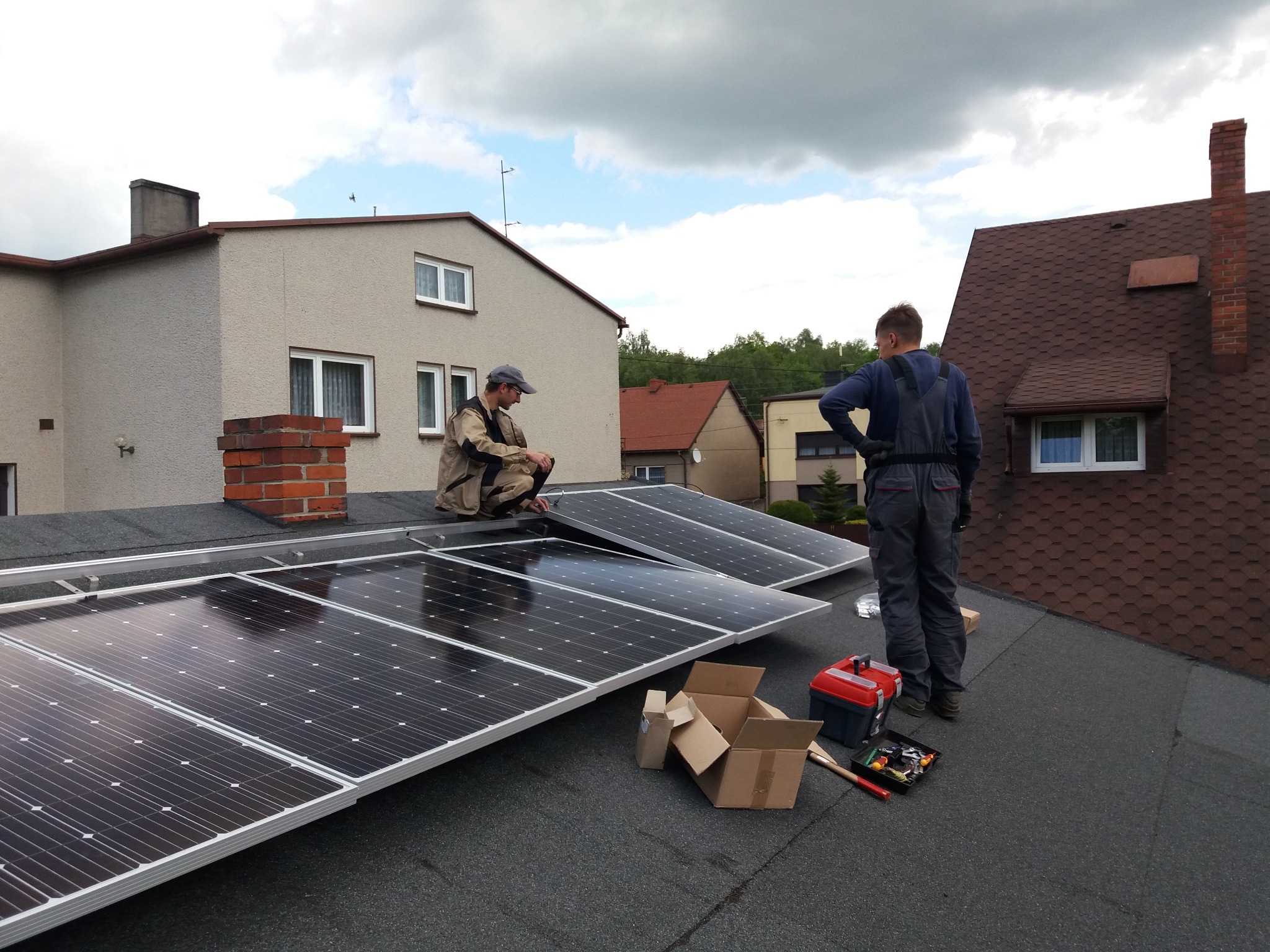 Montaż paneli słonecznych na dachu domu jednorodzinnego przez dwóch instalatorów. Widoczna skrzynka z narzędziami, kartony po panelach oraz fragmenty innych budynków w tle.
