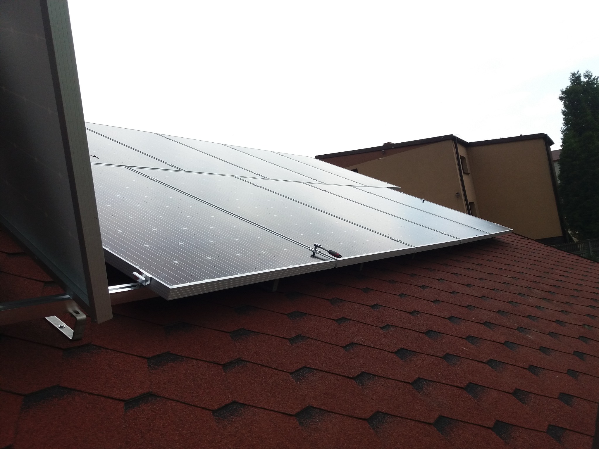 Panele słoneczne zamontowane na dachu pokrytym czerwoną dachówką bitumiczną, widoczny klucz nasadowy na jednym z paneli, budynek w tle.