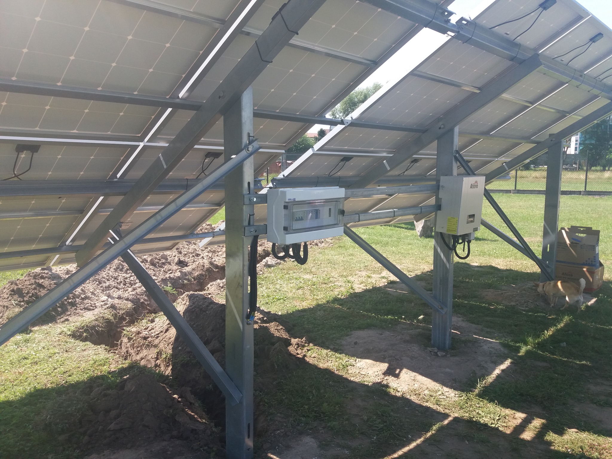 Widok od spodu na konstrukcję wsporczą paneli słonecznych z zamontowaną skrzynką elektryczną i falownikiem, świeżo po montażu z widocznymi wykopami w trawie, w tle kartony i pies.