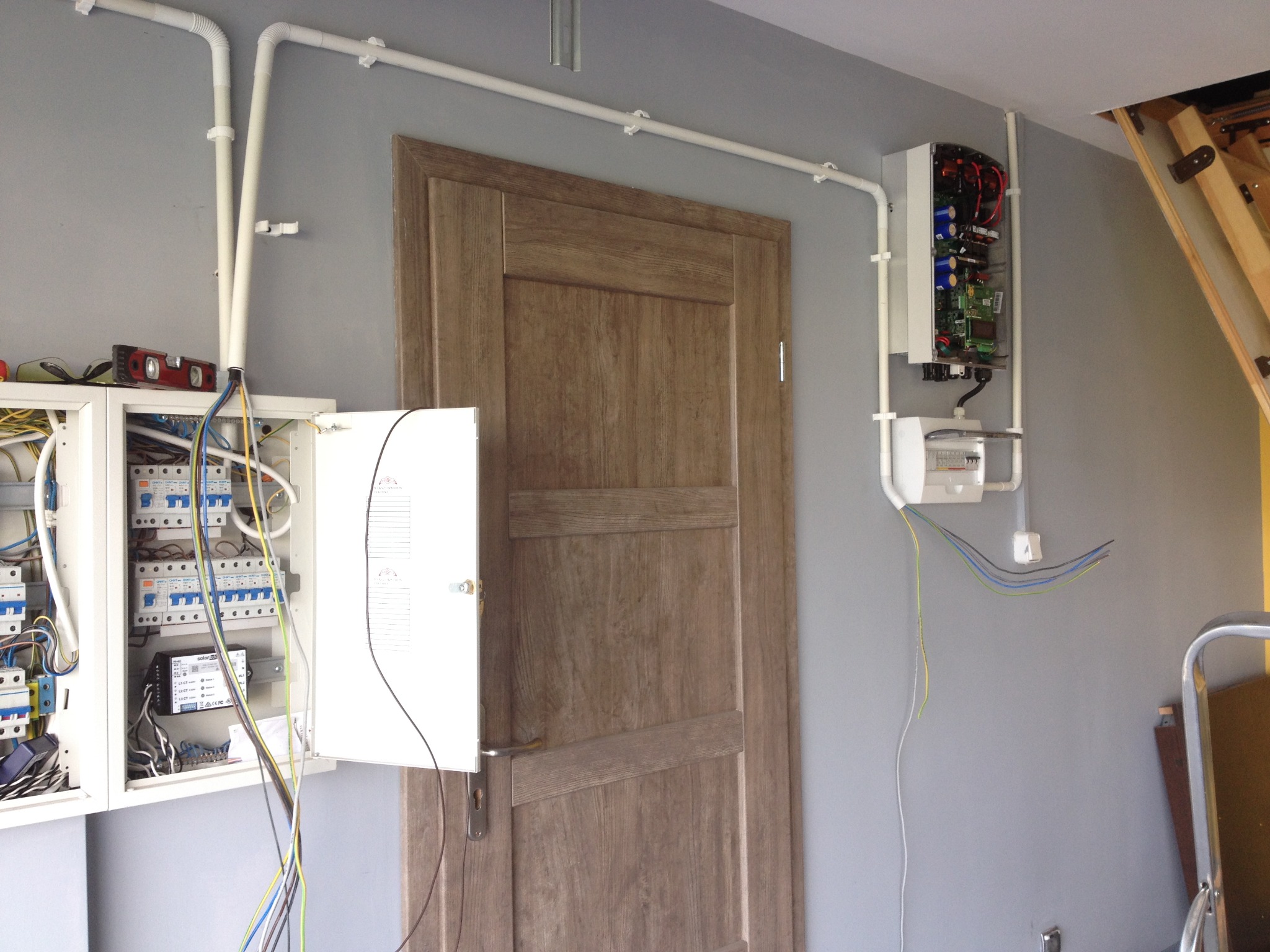 Instalacja elektryczna w szarej ścianie, z widocznymi rozdzielnicami, przewodami w białych rurkach PCV, poziomica na rozdzielnicy i drewniane schody strychowe w tle.