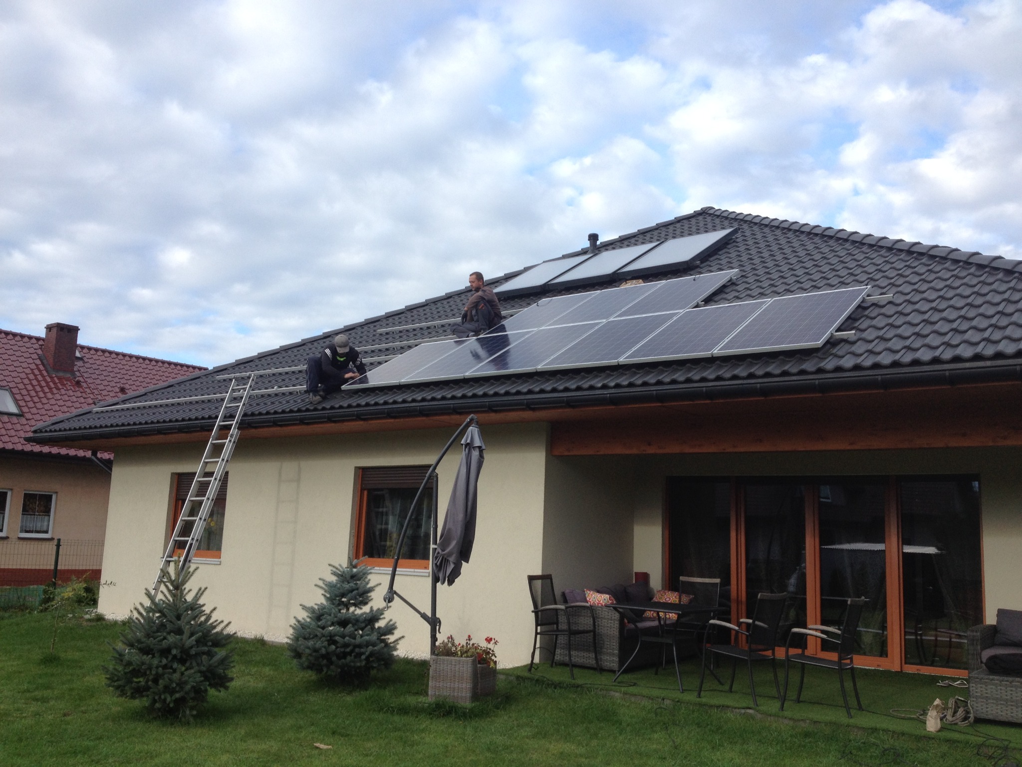 Montaż paneli słonecznych na dachu domu jednorodzinnego, widoczni dwaj instalatorzy pracujący na dachu, drabina oparta o ścianę, w tle sąsiedni dom i pochmurne niebo.