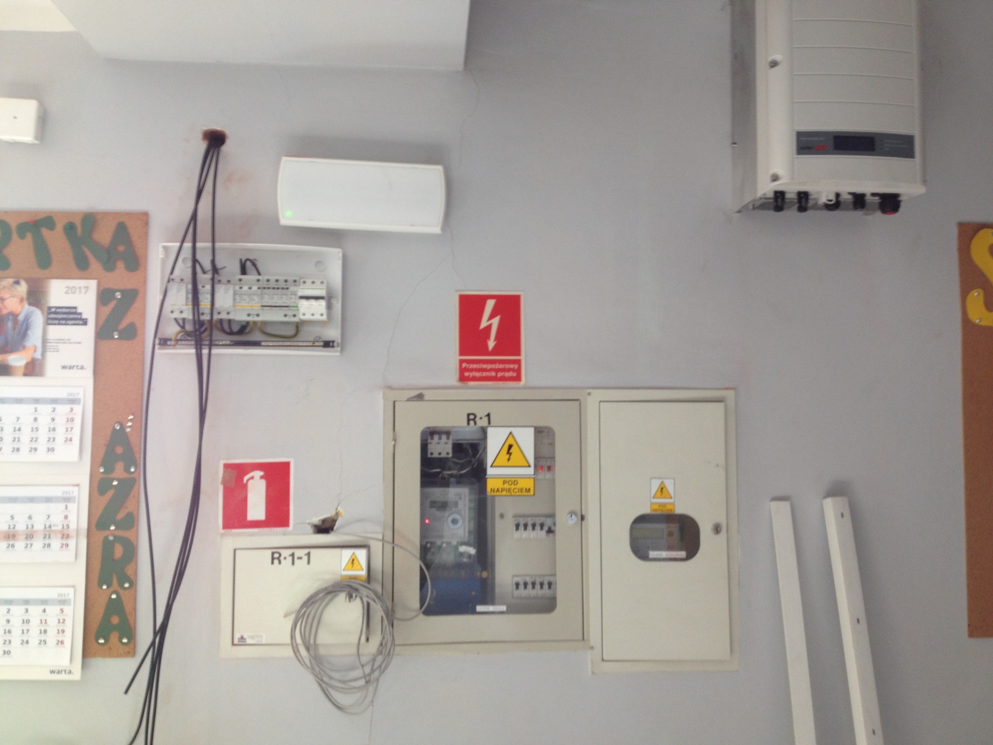 Szara ściana z zamontowaną białą skrzynką elektryczną, rozdzielnicą z bezpiecznikami, falownikiem fotowoltaicznym, kable wystające ze ściany, tabliczka ostrzegawcza o napięciu i kalendarz w tle.