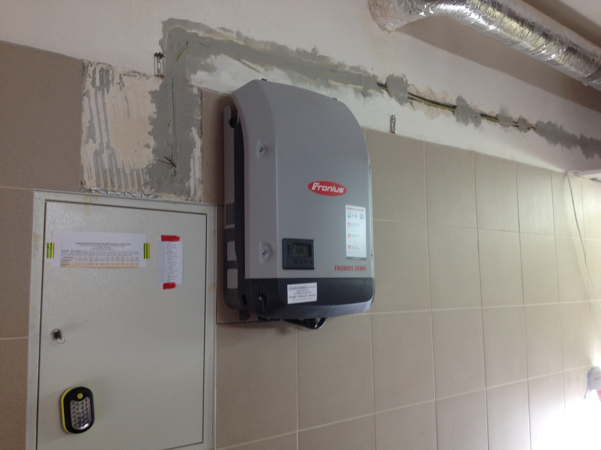 Szary falownik Fronius Symo zamontowany na kafelkowanej ścianie, widoczne kable w bruzdach pod sufitem i skrzynka elektryczna z latarką obok.