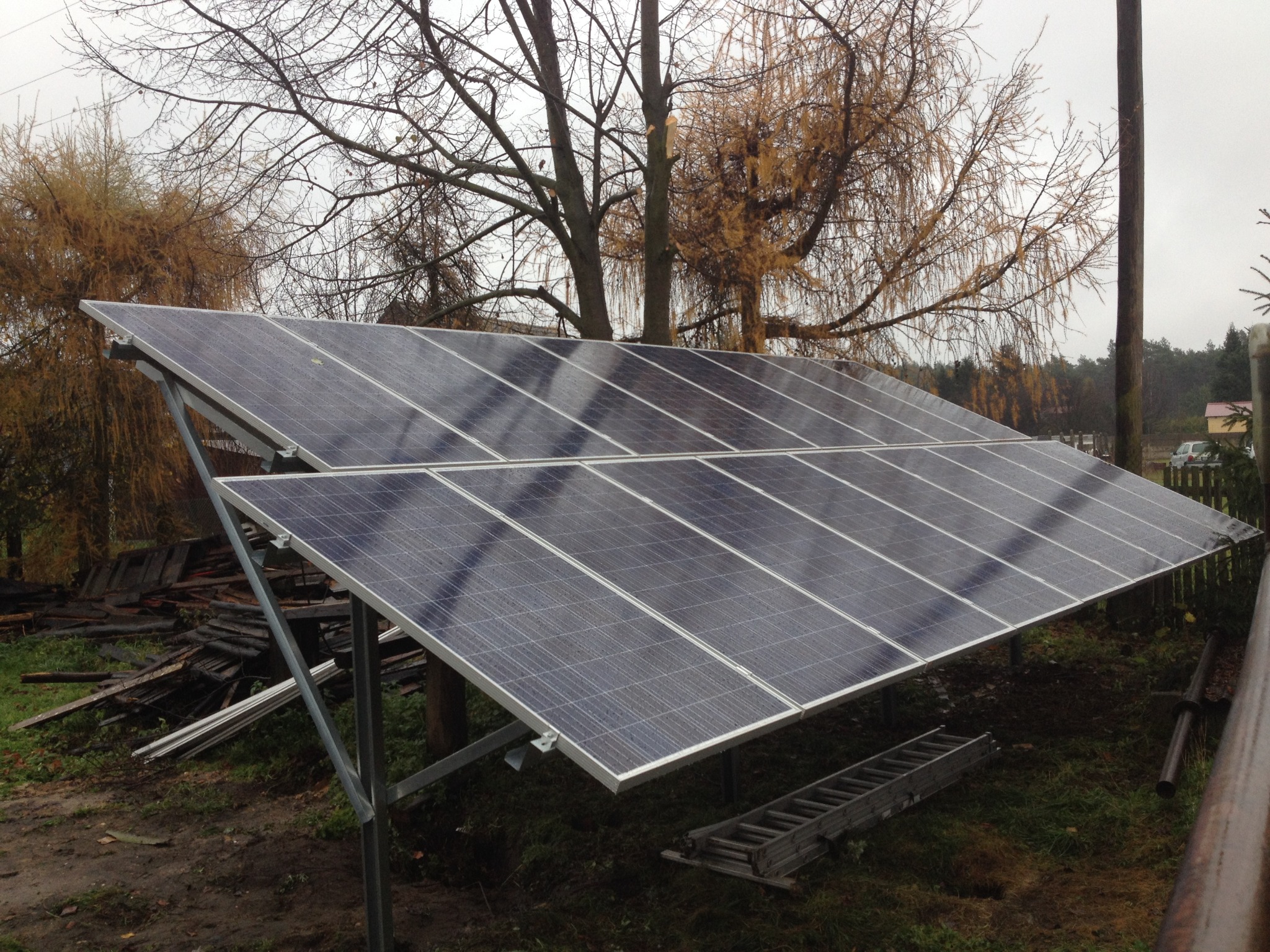 Panel fotowoltaiczny na metalowej konstrukcji w jesiennej scenerii, z widoczną stertą desek i drabiną w tle, w pochmurny dzień.