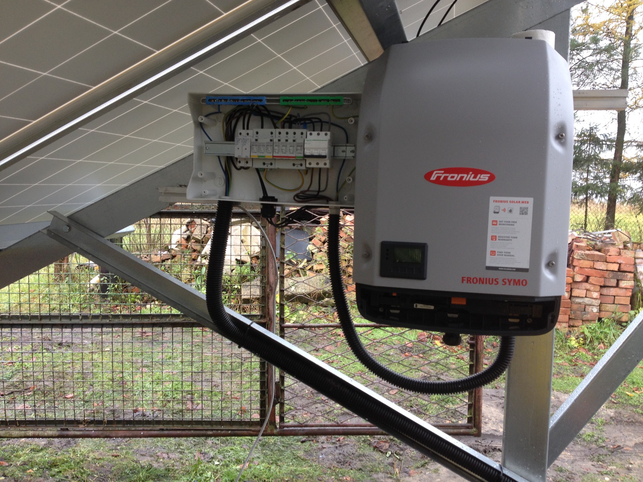 Szarej obudowy falownika Fronius Symo, zamontowanej pod panelami słonecznymi, z otwartą skrzynką elektryczną i widocznymi przewodami, na tle metalowej bramki i sterty cegieł.