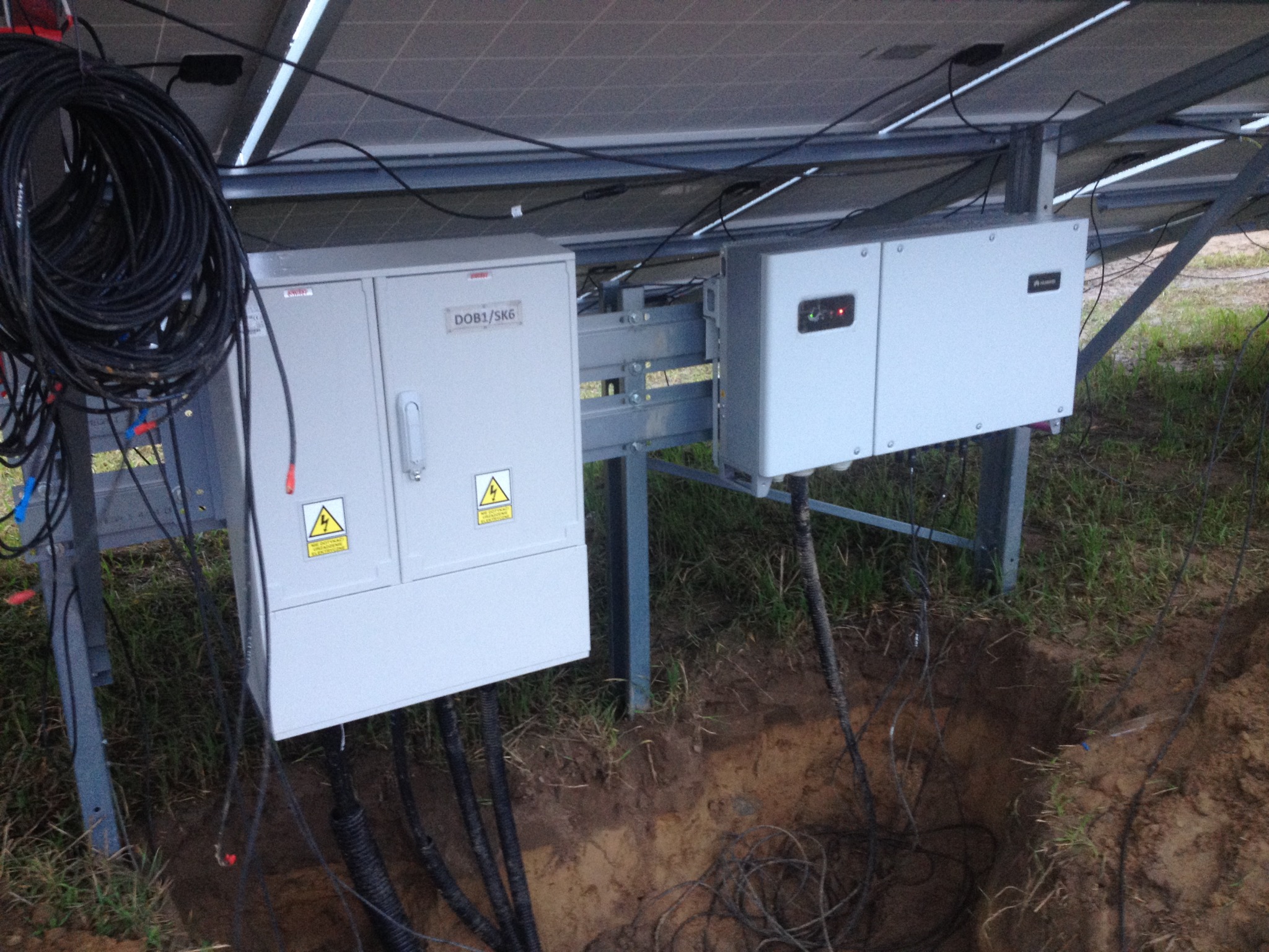 Panele słoneczne z podłączonymi skrzynkami elektrycznymi i kablem zwisającym z konstrukcji; widoczny wykop w ziemi.