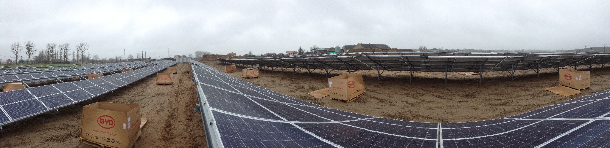 Rozległa farma fotowoltaiczna w trakcie instalacji, widoczne rzędy paneli słonecznych i kartony BYD na paletach, pochmurne niebo w tle.
