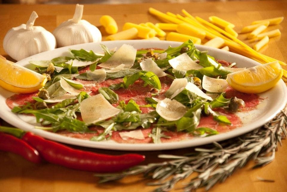 Carpaccio wołowe z rukolą, parmezanem, kaparami i cytryną, podane na białym talerzu z czosnkiem, papryczką chili i rozmarynem w tle.