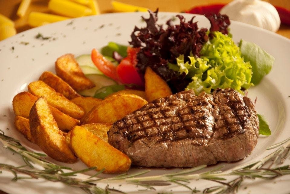 Grillowany stek wołowy z ziemniakami w łupinach, sałatką i gałązką rozmarynu na białym talerzu.