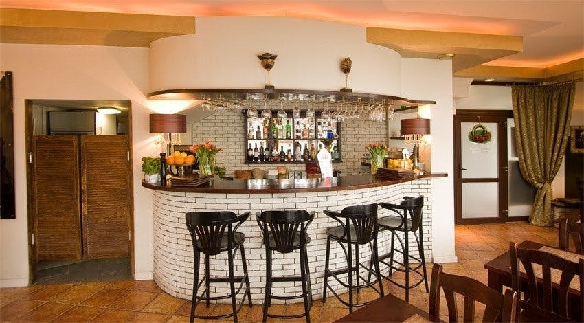 Elegancki bar z białej cegły z czarnymi stołkami, za nim półki z alkoholem i szklankami, dekoracje z kwiatów i owoców, w tle drewniane drzwi i zasłony.