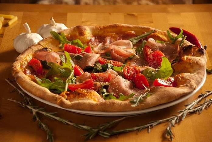 Pizza z szynką parmeńską, pomidorami i rukolą na cienkim cieście, podana na białym talerzu z dekoracją z gałązek rozmarynu i główkami czosnku w tle.
