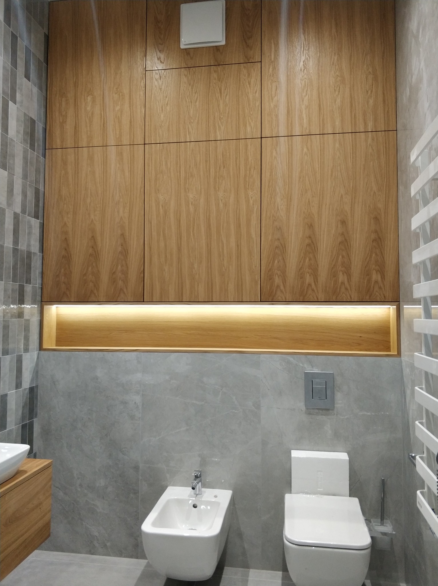 Nowoczesna łazienka z drewnianą zabudową szafek podświetlonych od dołu, szare płytki imitujące marmur, biała ceramika sanitarna i grzejnik drabinkowy.