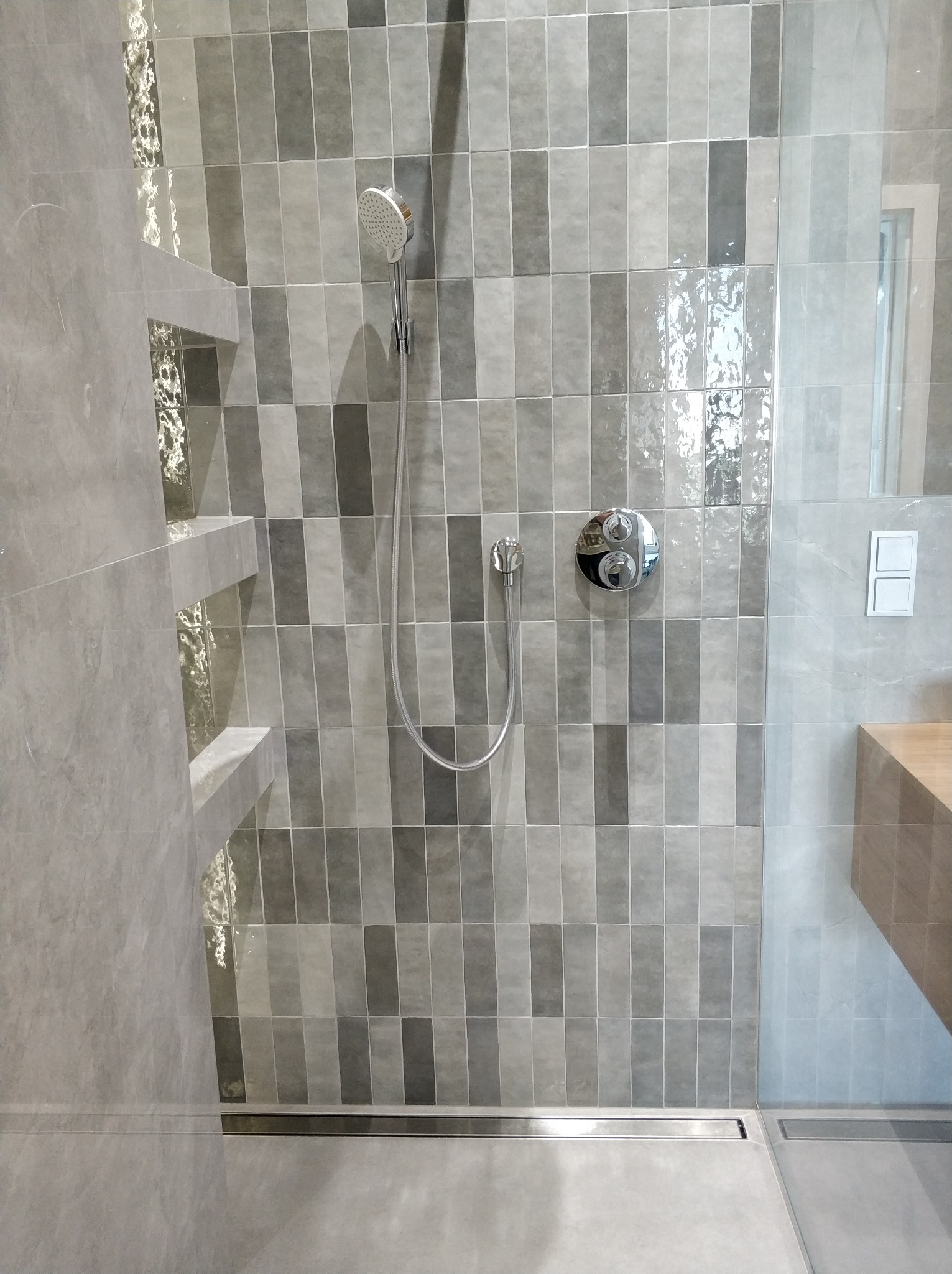 Wykończona kabina prysznicowa z szarymi, pionowo ułożonymi płytkami o zróżnicowanym odcieniu, chromowaną armaturą i odpływem liniowym w podłodze, widoczny fragment drewnianego blatu umywalki.