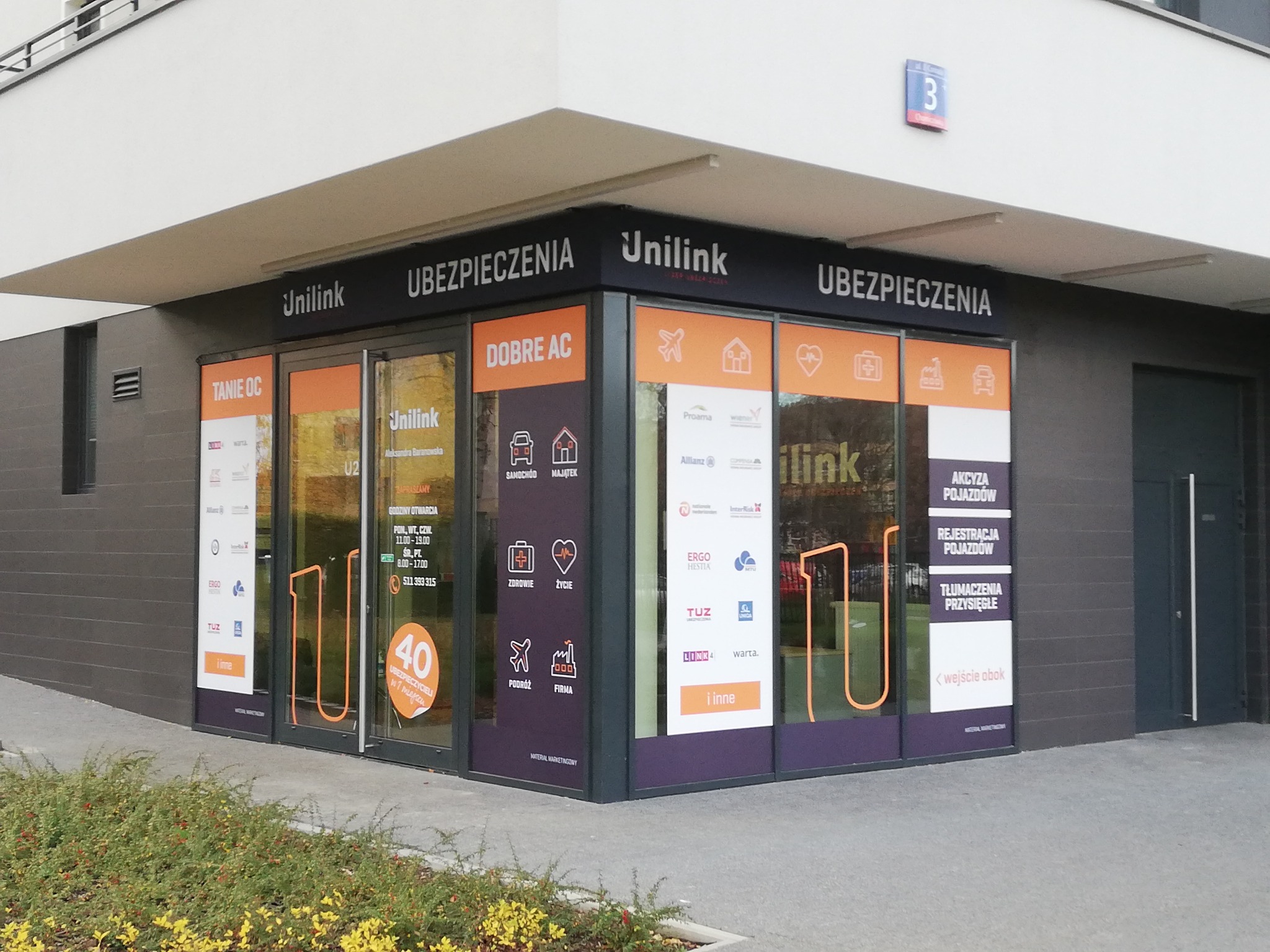 Fasada punktu stacjonarnego Unilink Ubezpieczenia z widocznymi naklejkami informacyjnymi na szybach dotyczącymi ubezpieczeń komunikacyjnych, majątkowych, na życie i zdrowotnych oraz podróżnych...