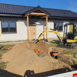 Żółta mini koparka Yanmar w trakcie prac ziemnych przy budowie domu z białych bloczków, widoczne wykopane rowy, rury kanalizacyjne, ubijarka, poziomica i tablica informacyjna budowy.