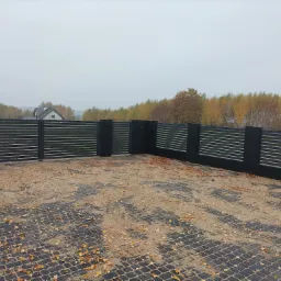 Nowoczesne, poziome ogrodzenie panelowe w kolorze antracytowym, z betonowymi słupkami, na posesji wyłożonej kostką brukową z widocznymi opadłymi liśćmi, w tle drzewa jesienne i dom.